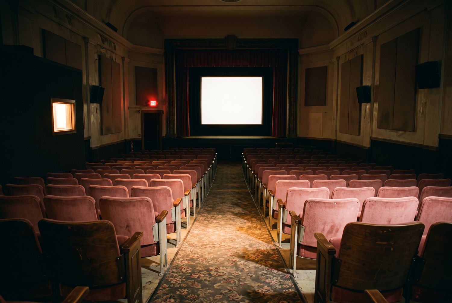 The house seen from the head of the centre aisle at the back wall: raked rows of rose seats running down to the lit screen, with the glowing projection port in the wall to the left.