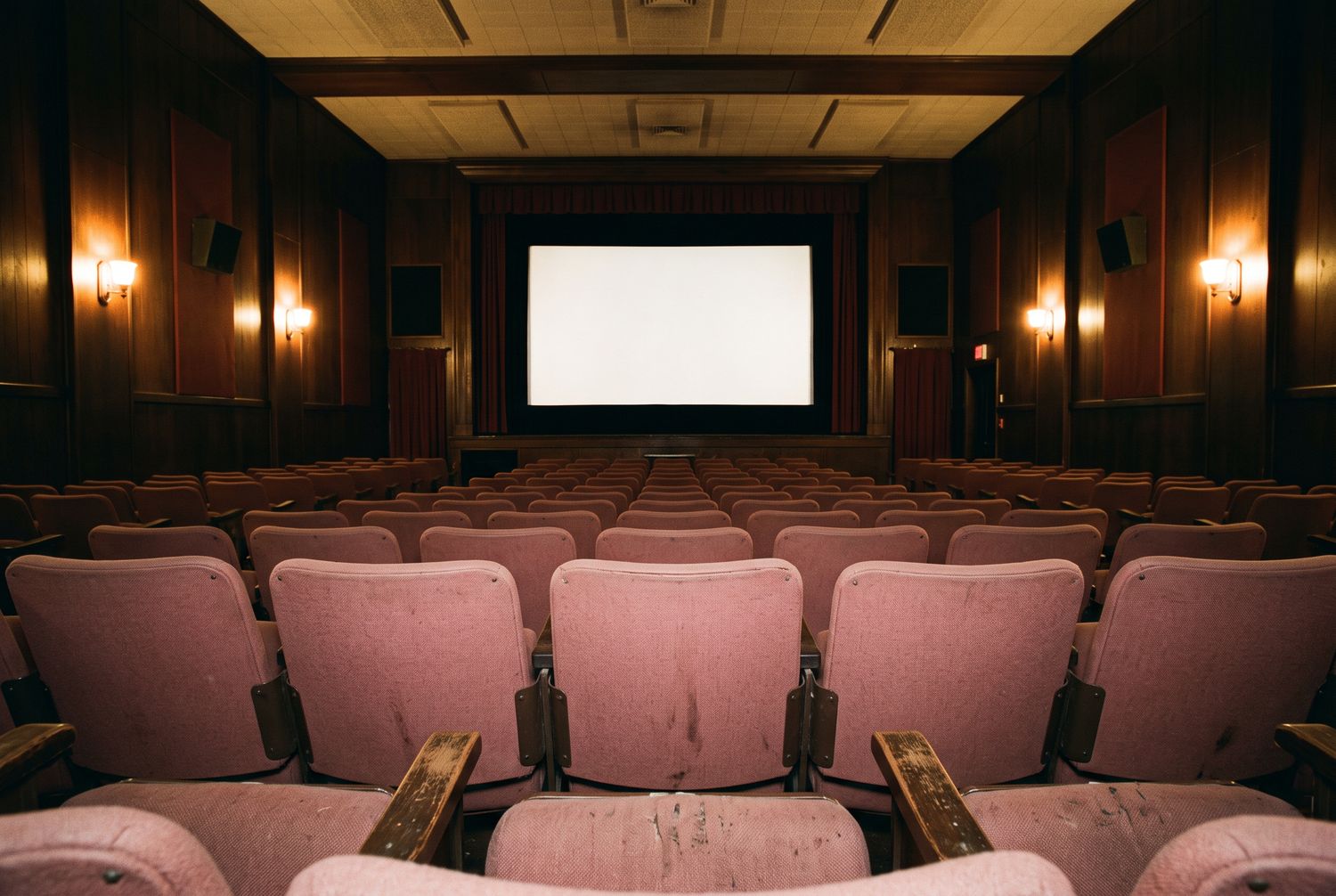 Rows of empty rose seats with worn wooden armrests seen at seated height from the middle of the house, the lit screen square and level ahead.
