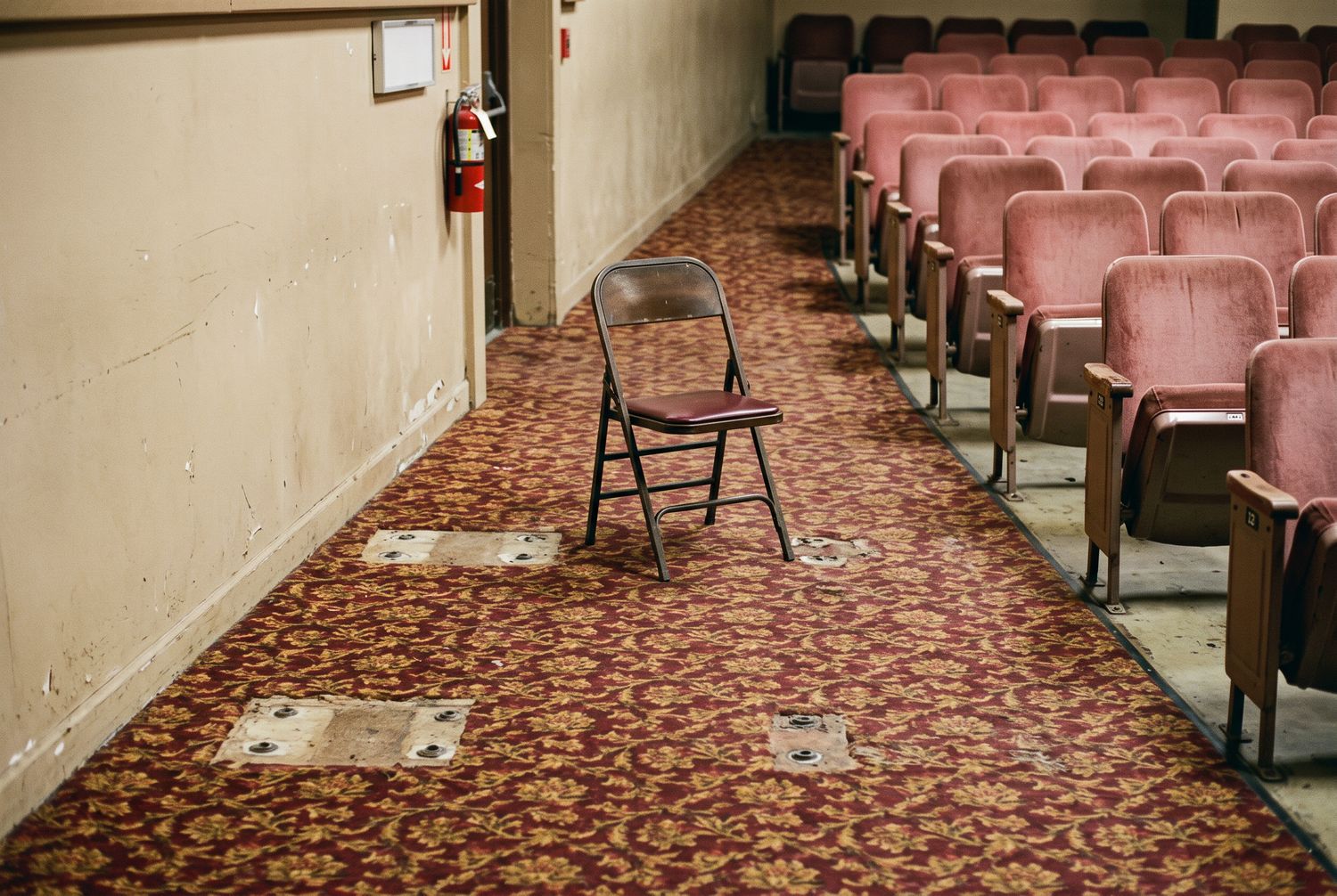 A cleared level area at the head of an aisle where fixed seats were removed, the capped floor bolts still visible in the carpet, one folding chair standing beside it and rose seats to the right.